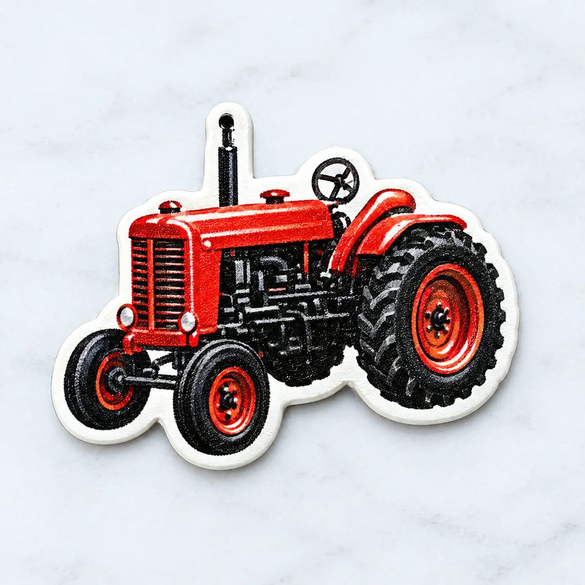 Nahaufnahme eines einzelnen Anhängers in Form eines roten Oldtimer-Traktors mit weißem Rand und kleinem Aufhängeloch, auf hellem Marmorhintergrund. 3