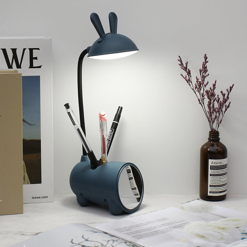 Dunkelblaue Hasenlampe mit Licht an, drei Stifte im Halter des Fußes, daneben Bücher und eine Vase mit Trockenblumen.