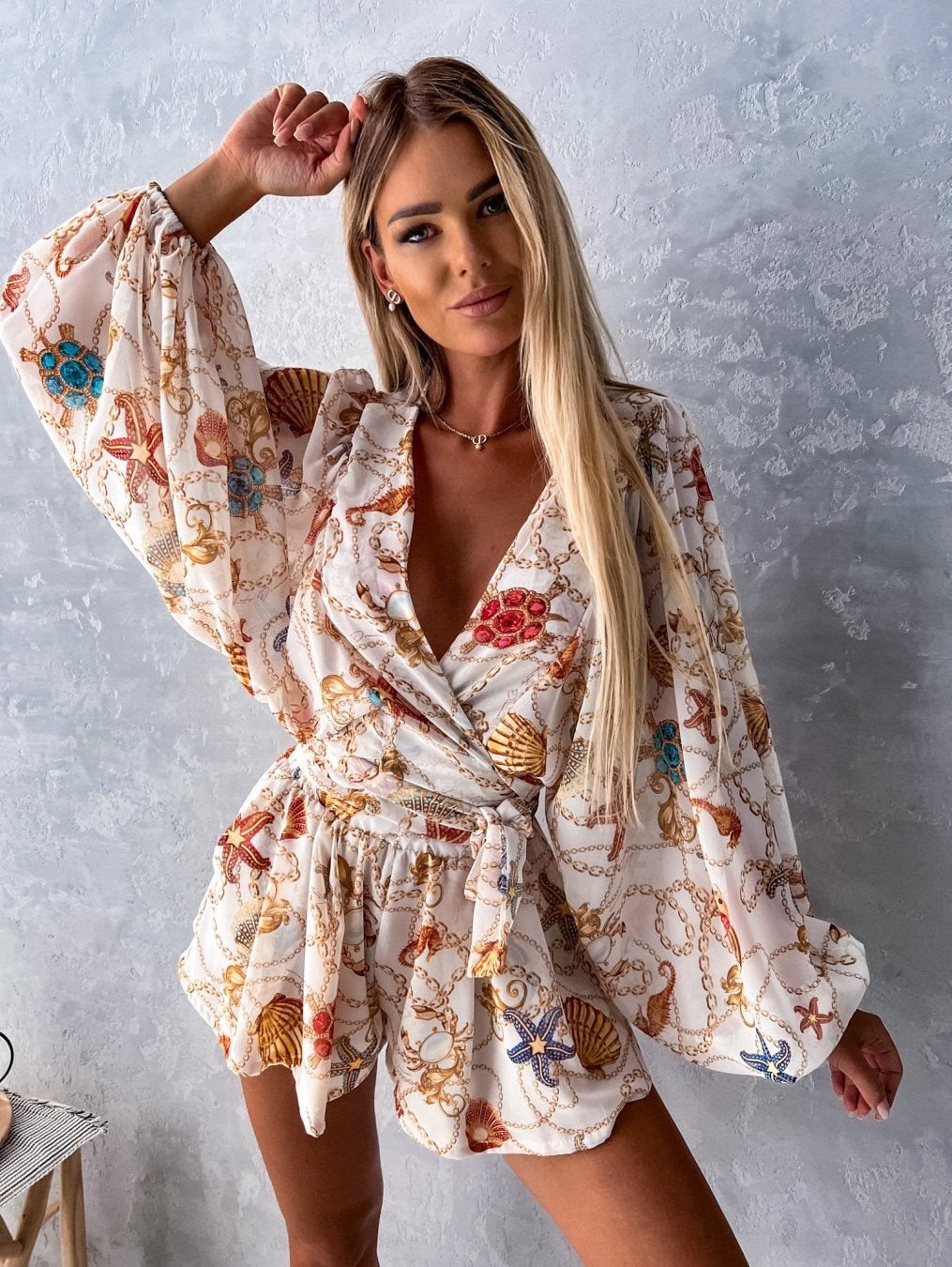 Frau in weißem, gemustertem Sommerkleid mit Muschel- und Kettenmotiven vor grauer Wand.