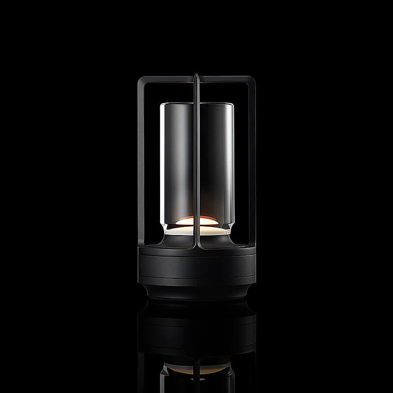 Schwarze Tischlampe mit Metallrahmen und Glaszylinder leuchtet warm vor schwarzem Hintergrund, unten gespiegelt. 11