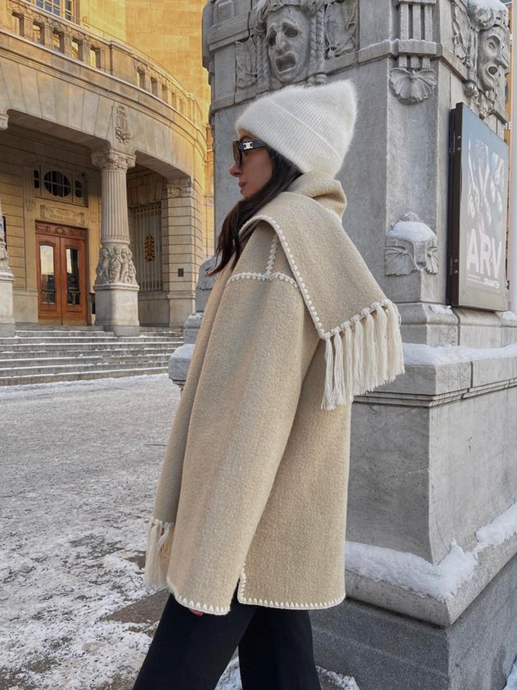 Frau in beiger Wolljacke mit Fransen, weiße Mütze, Sonnenbrille, Wintermode, Stadtumgebung.