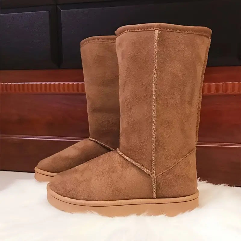 Braune Wildleder-Stiefel, kniehoch, auf weißem Teppich vor Holzwand.