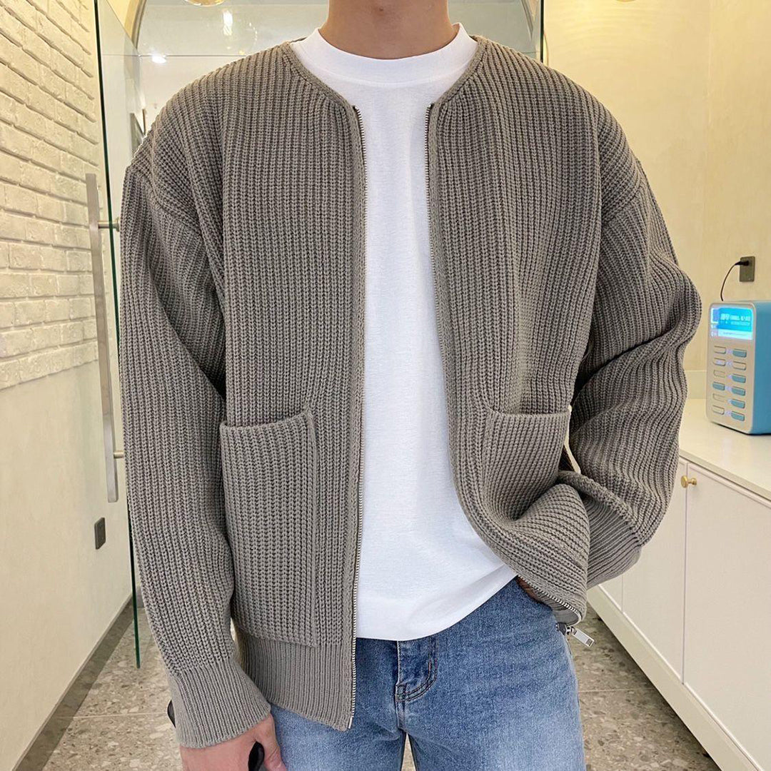 Mann in khakifarbener Strickjacke, weißem T-Shirt und blauer Jeans in modernem Raum.