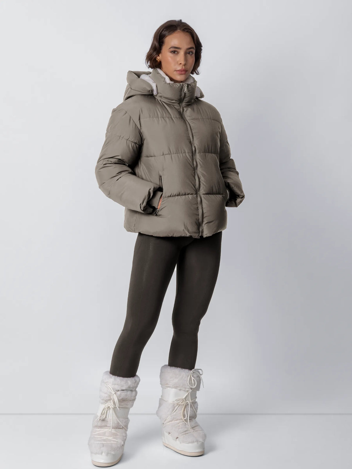 Ganzkörperansicht: Model in taupefarbener Steppjacke mit Kapuze, dunklen Leggings und weißen Plüschstiefeln, Hände in den 21