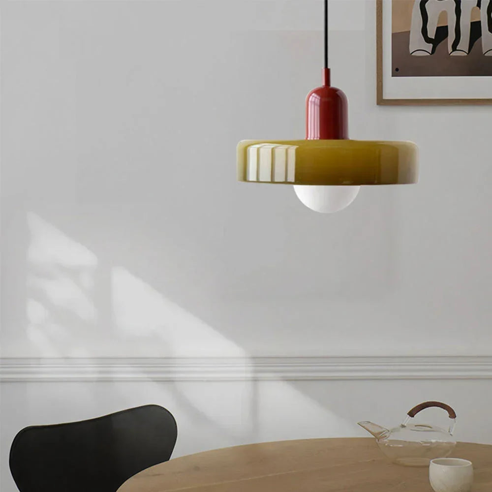 Pendelleuchte mit rotbrauner Kappe über einem Esstisch, im Hintergrund eine helle Wand mit Bilderrahmen und eine Glaskanne. 25