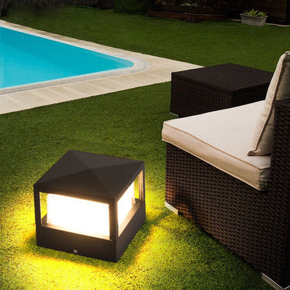Quadratische Leuchte mit Pyramidendach, leuchtend auf dem Rasen neben einem Pool und einem Rattan-Sofa.