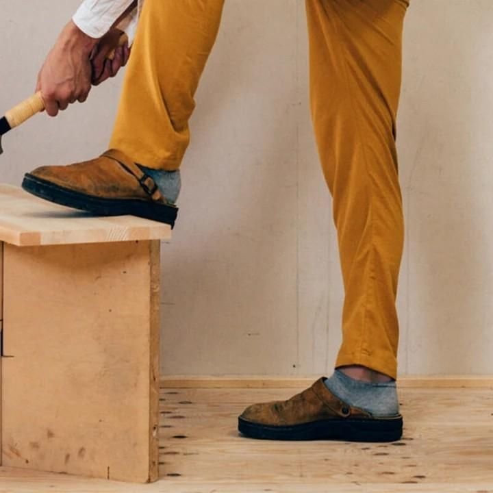 Person in senffarbener Hose und grauen Socken trägt die braunen Clogs, ein Fuß steht auf einer Holzkiste 3