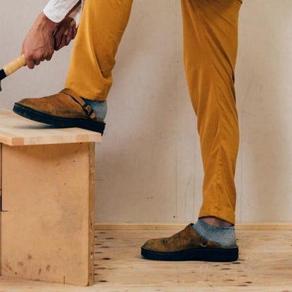 Person in senffarbener Hose und grauen Socken trägt die braunen Clogs, ein Fuß steht auf einer Holzkiste 3