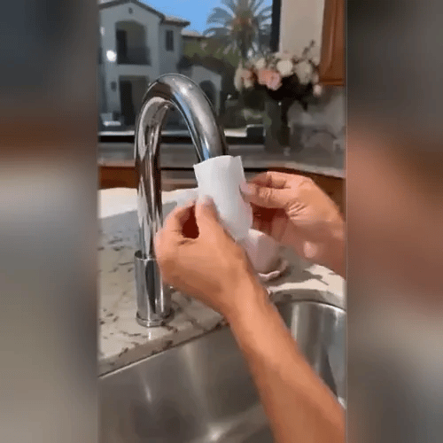 Zwei Hände halten einen weißen Filterbeutel an einen verchromten Wasserhahn über einer Küchenspüle, Anbringen des Filters. 2
