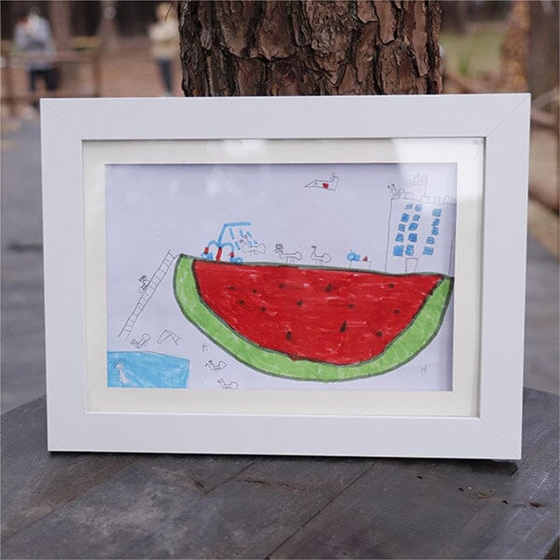 Weißer Bilderrahmen im Querformat mit einer Kinderzeichnung einer riesigen Wassermelone, im Freien auf einem Holztisch vor einem Baumstamm aufgestellt. 21