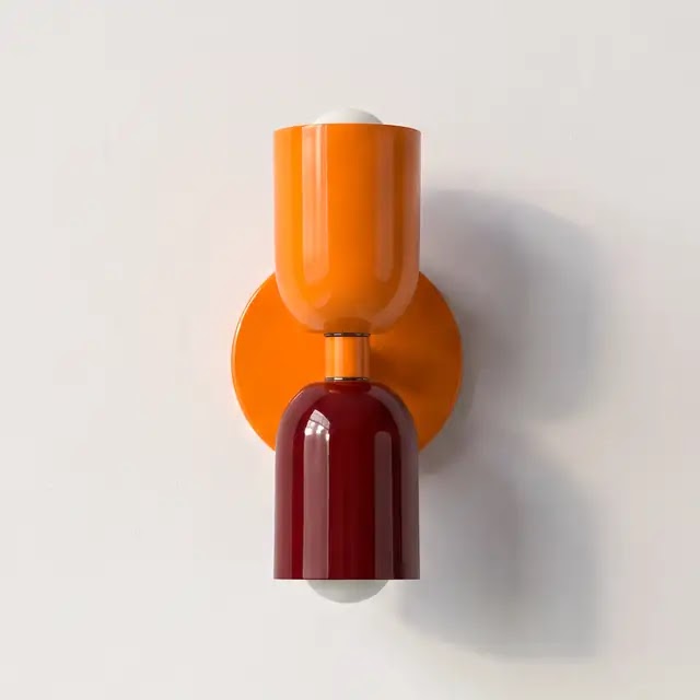 Wandlampe mit leuchtend orangem oberem Schirm und dunkelrotem unterem Schirm an einer orangefarbenen runden Wandplatte, frontal vor heller Wand. 25