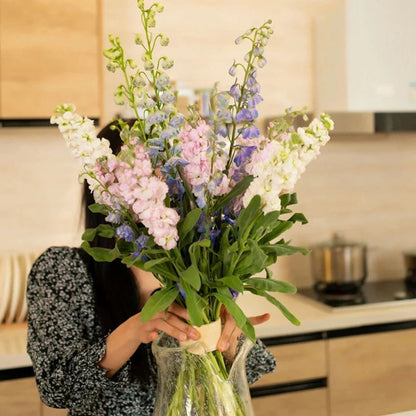 Frau in gemustertem Oberteil hält in der Küche einen aufrechten Strauß aus rosa, weißen und blauen Blumen in einer Glasvase. 2