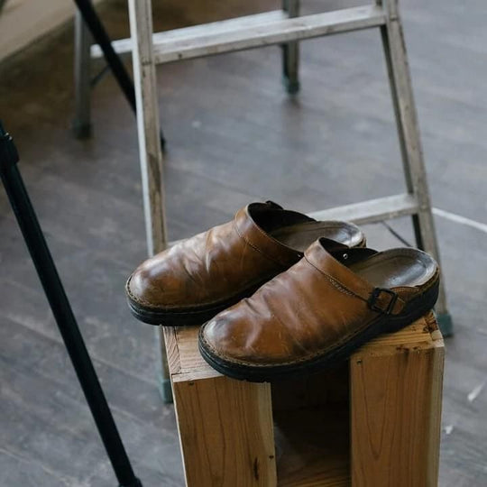 Ein Paar abgetragene braune Leder-Clogs mit Schnalle auf einem Holzhocker vor einer Leiter 1