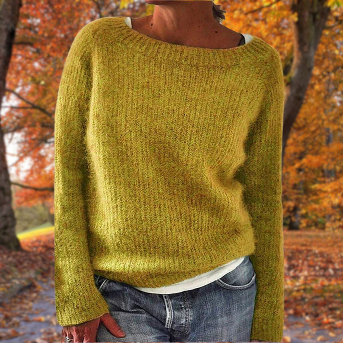 Senfgelber Strickpullover mit rundem Ausschnitt, getragen von einer Frau mit Jeans und weißem Shirt vor herbstlichem Laub. 1