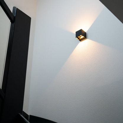 Schwarze Würfel-Wandlampe im Treppenhaus an weißer Wand, Blick von unten, mit warmem Lichtkegel nach oben und unten. 2