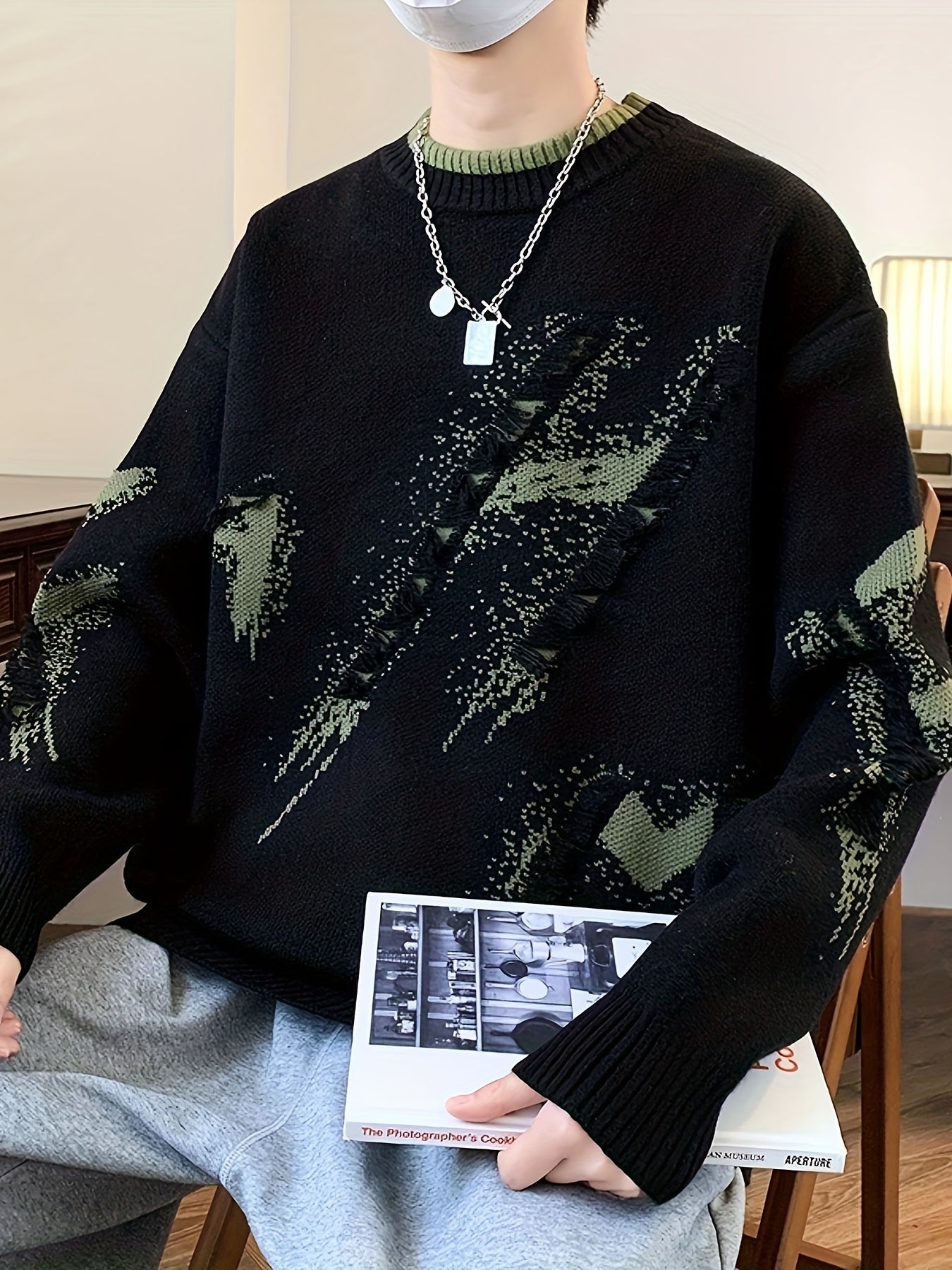 Mann in schwarzem Strickpullover mit grünem Muster, silberner Kette, hält ein Buch.