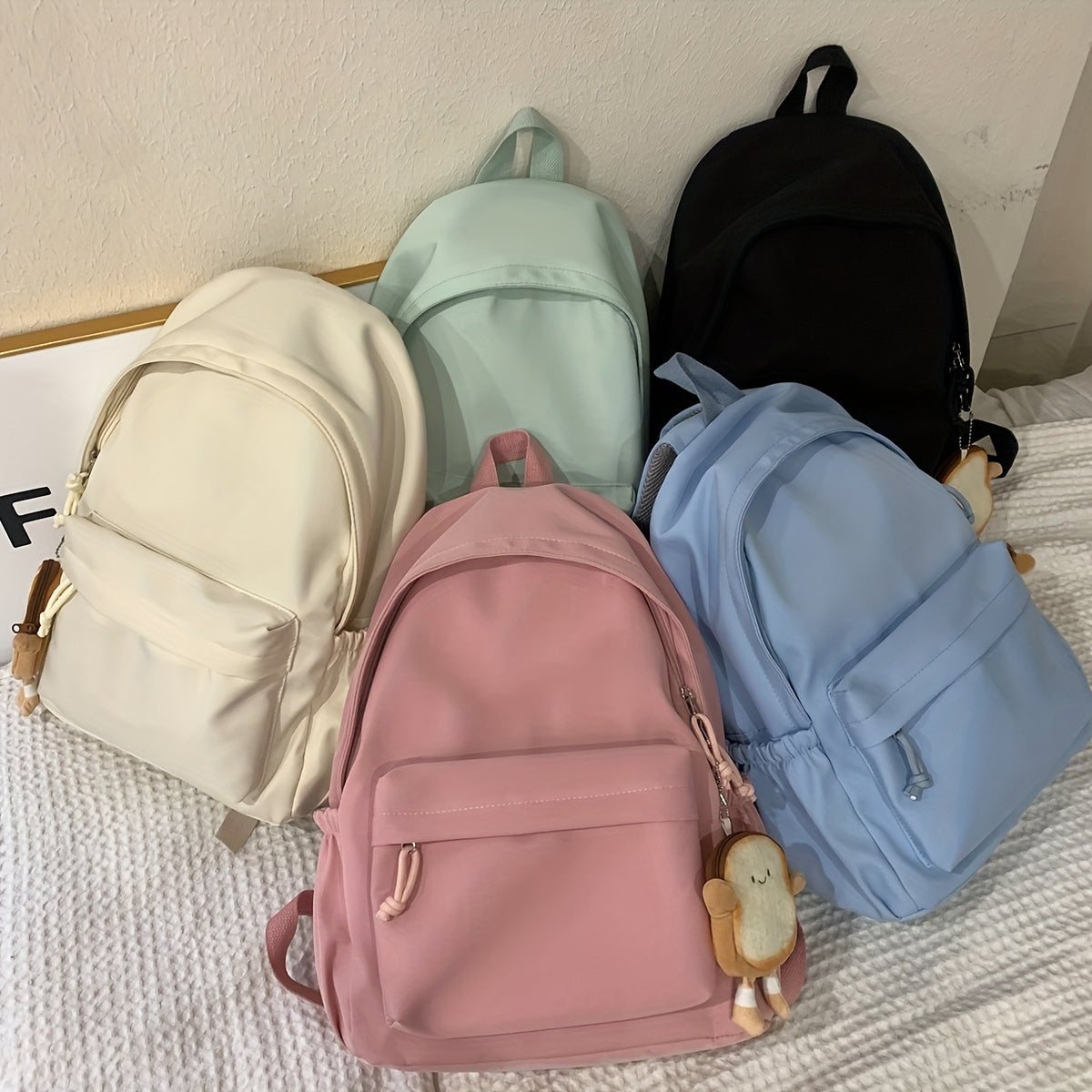 Fünf Rucksäcke in Creme, Mint, Schwarz, Rosa und Hellblau liegen nebeneinander auf einem weißen Bett, daran ein kleiner Brot-Anhänger.