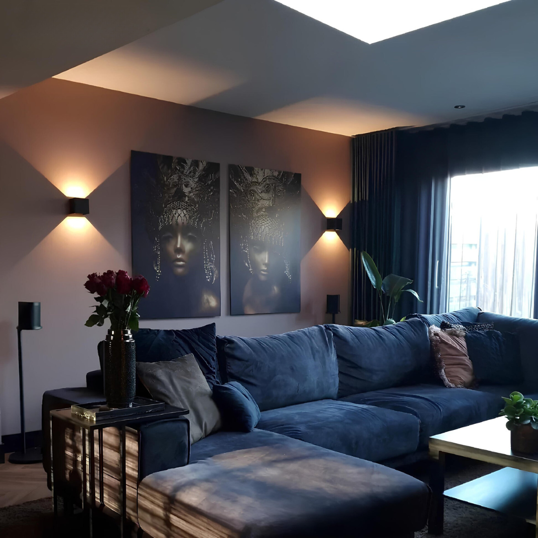Zwei schwarze Wandlampen über einem blauen Samtsofa im Wohnzimmer, zwischen zwei Wandbildern, mit warmem Licht. 9