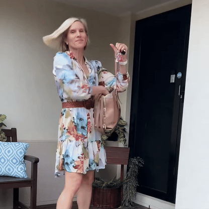 Frau mit glatten blonden Haaren in geblümtem Kleid schwingt ihr Haar vor einer Haustür, Handtasche und Schlüssel in der Hand. 8