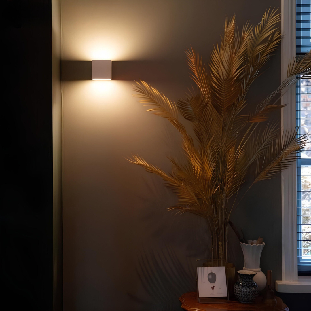 Weiße Würfel-Wandlampe an dunkler Wand mit warmem Licht, daneben große goldene Palmwedel in einer Vase. 4