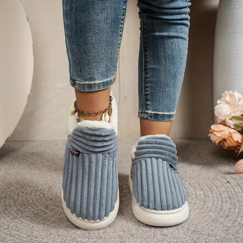 Graublaue gerippte Hausschuhe von vorn an den Füßen einer Person in Jeans mit Muschel-Fußkettchen.