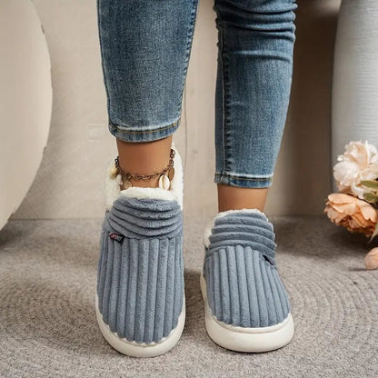 Graublaue gerippte Hausschuhe von vorn an den Füßen einer Person in Jeans mit Muschel-Fußkettchen.