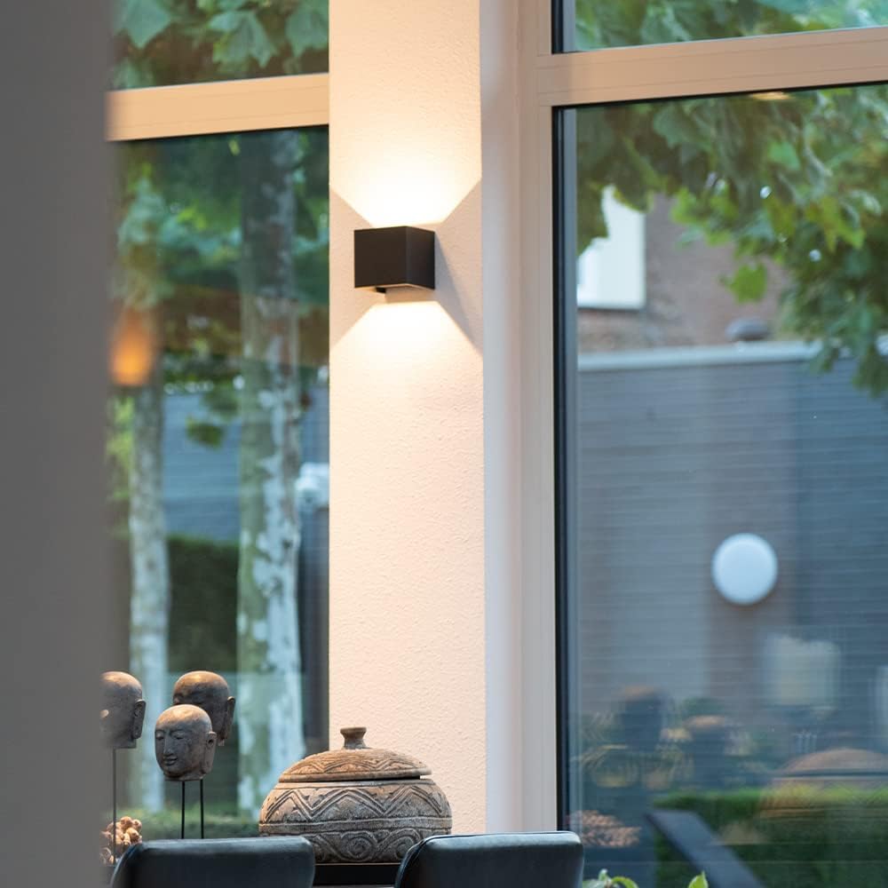 Schwarze Würfel-Wandlampe an einer weißen Wandsäule zwischen Fensterscheiben, davor Dekoköpfe und eine verzierte Schale. 8