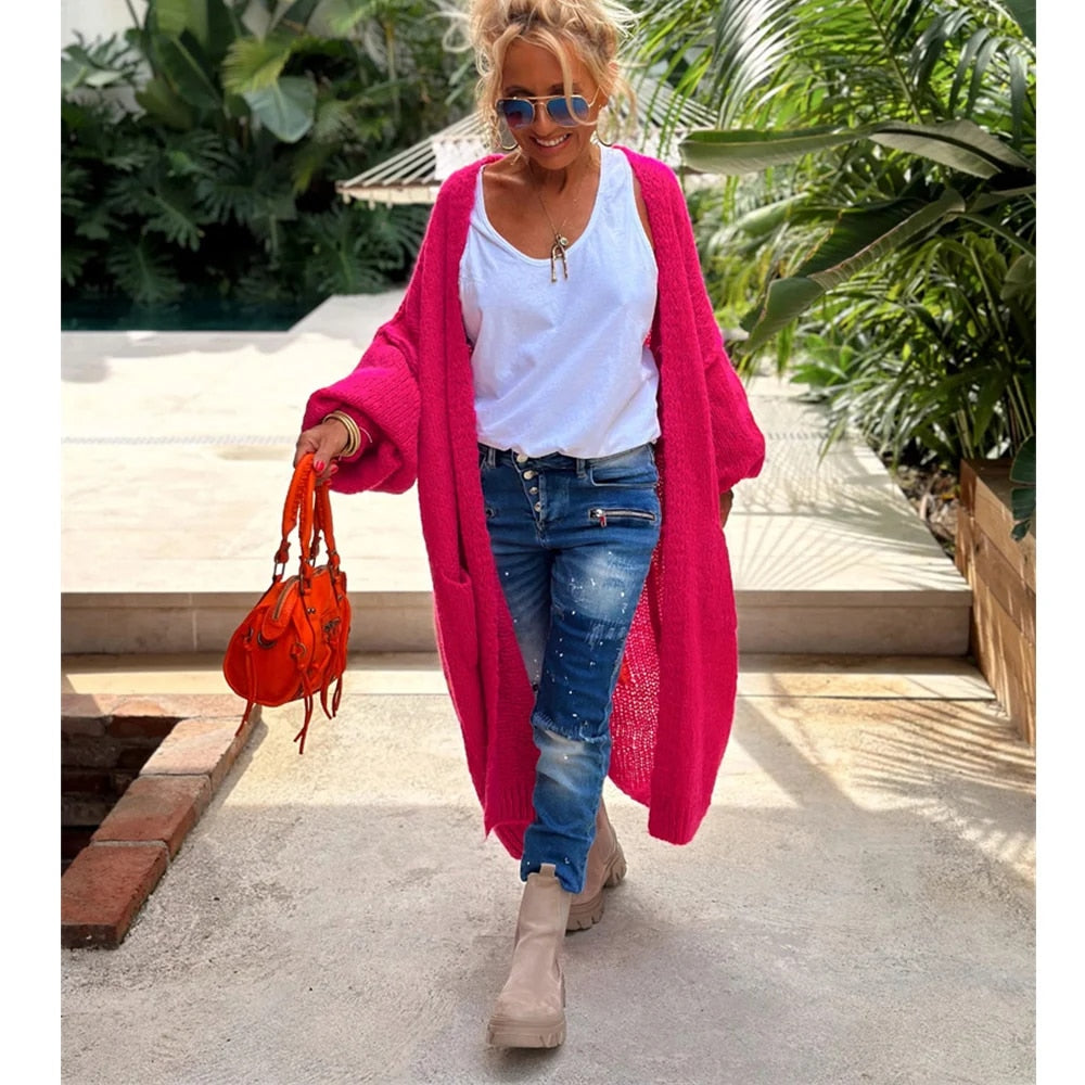Frau in pinkem Strick-Cardigan, weißem T-Shirt, Jeans und Stiefeln, hält orange Tasche.