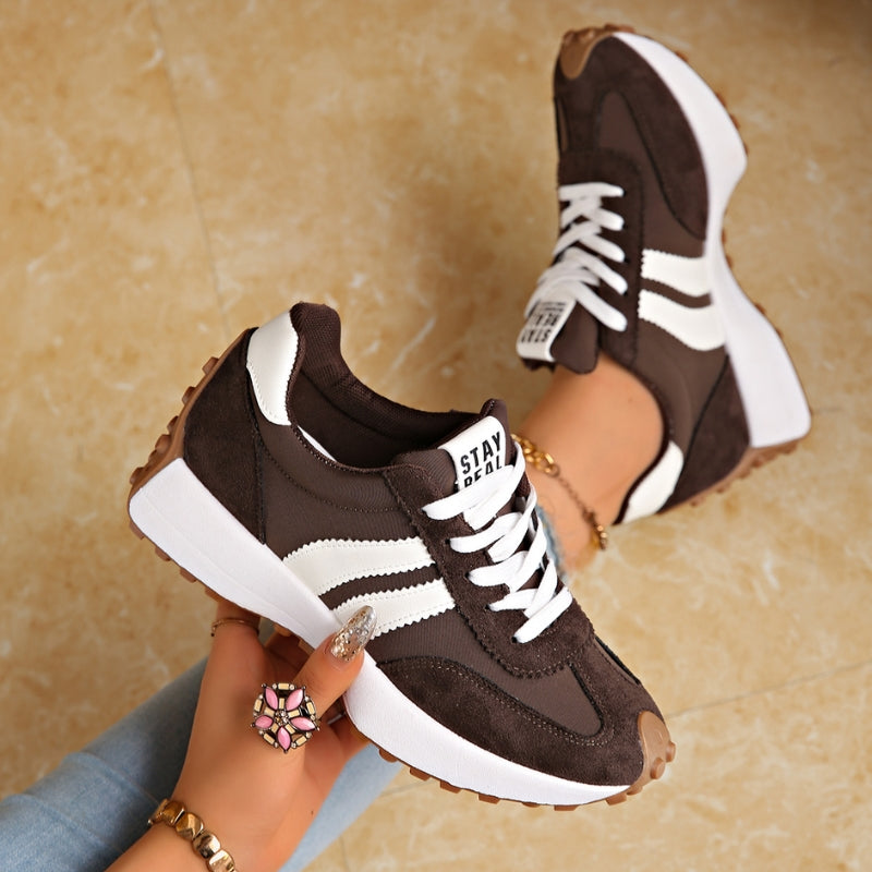 Dunkelbraune Sneaker mit weißen Streifen und Plateausohle, in der Hand gehalten, Fuß mit Armband im Hintergrund 3