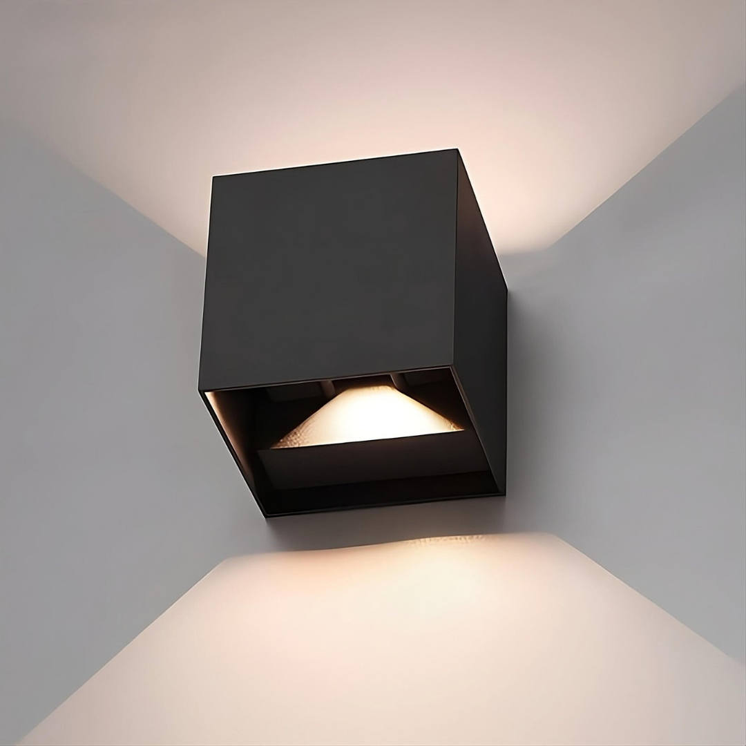 Nahaufnahme einer schwarzen Würfel-Wandlampe mit sichtbarer LED-Öffnung unten, Licht fällt nach oben und unten auf graue Wand. 16