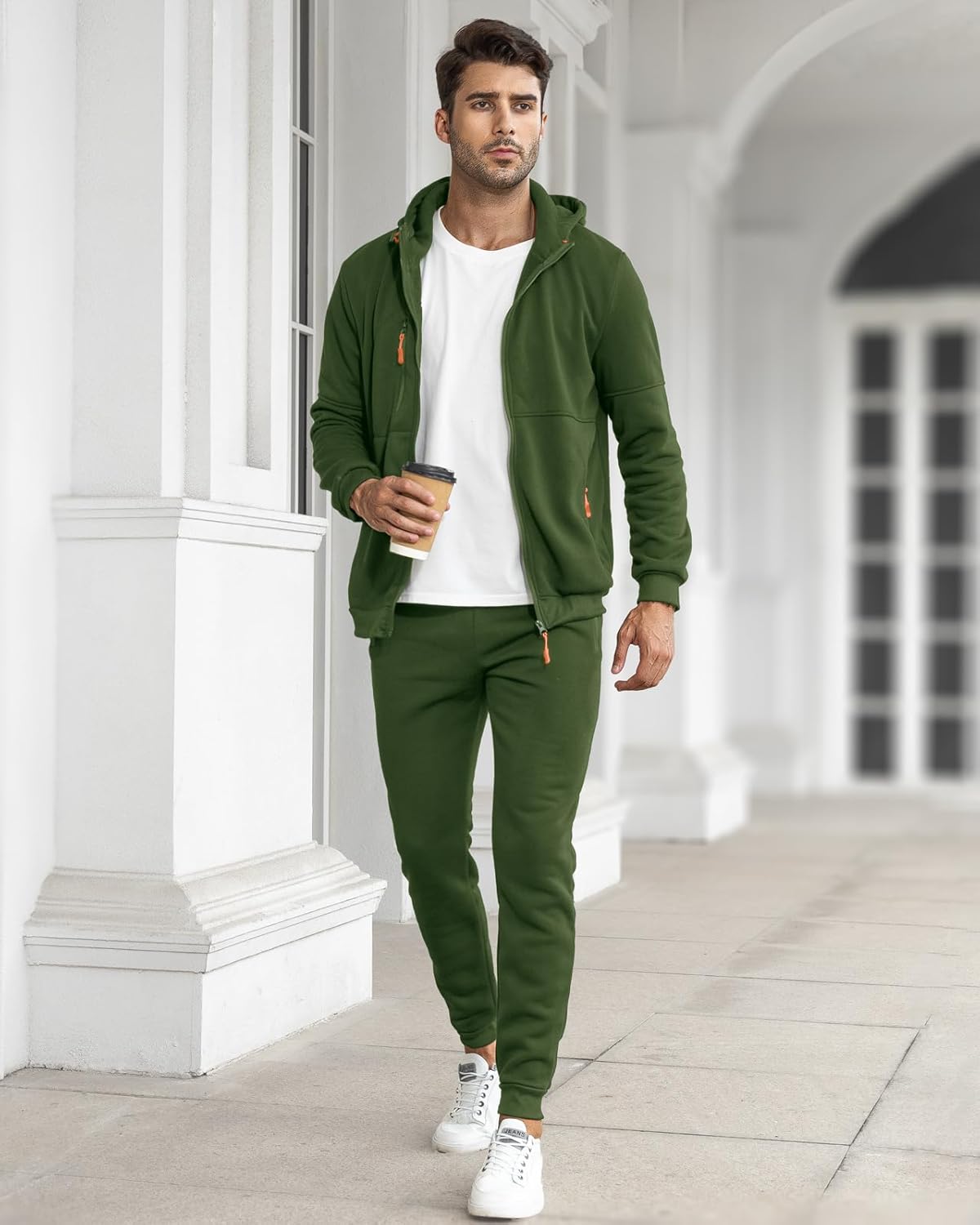 Mann in grünem Jogginganzug mit Kapuze, hält Kaffeebecher, weiße Sneaker, urbaner Hintergrund.
