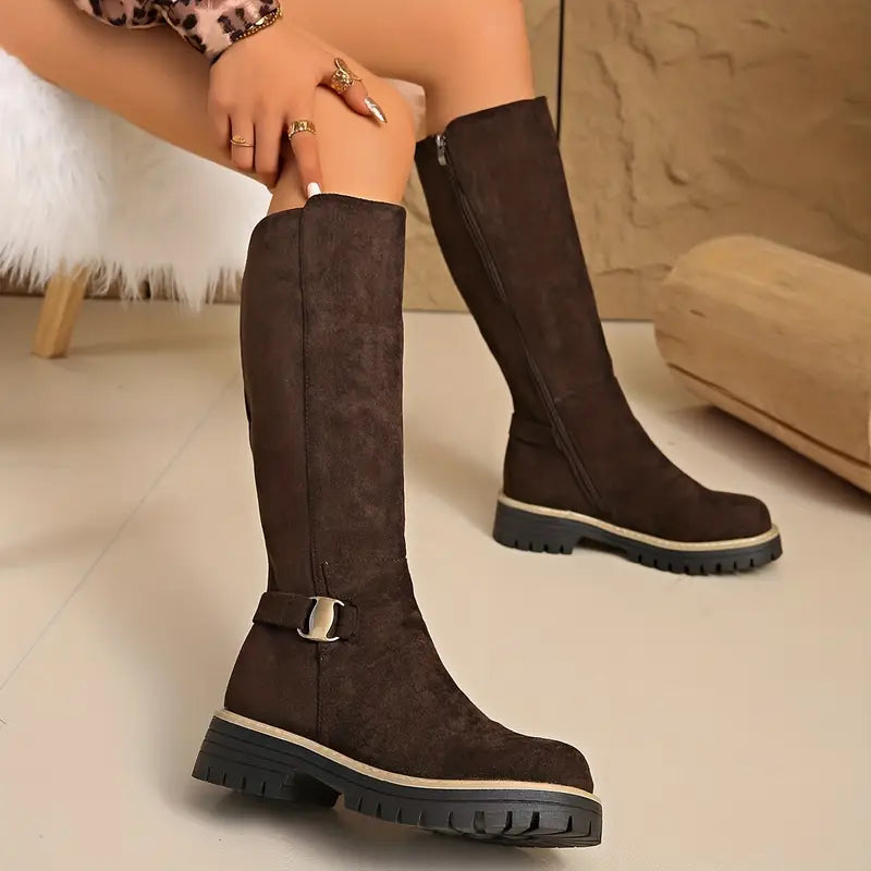 Braune kniehohe Stiefel im Sitzen, mit Hand am Schaft und dem zweiten Stiefel im Hintergrund.