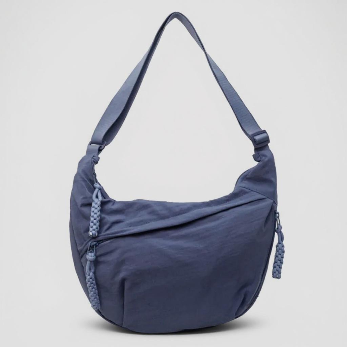 Blaue halbmondförmige Nylontasche mit breitem verstellbarem Riemen und geflochtenen Reißverschlussanhängern, frontal vor hellgrauem Hintergrund