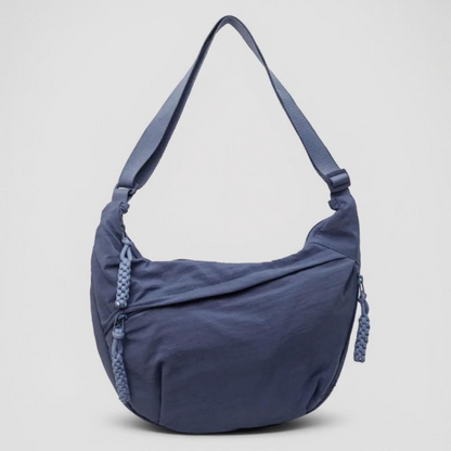 Blaue halbmondförmige Nylontasche mit breitem verstellbarem Riemen und geflochtenen Reißverschlussanhängern, frontal vor hellgrauem Hintergrund