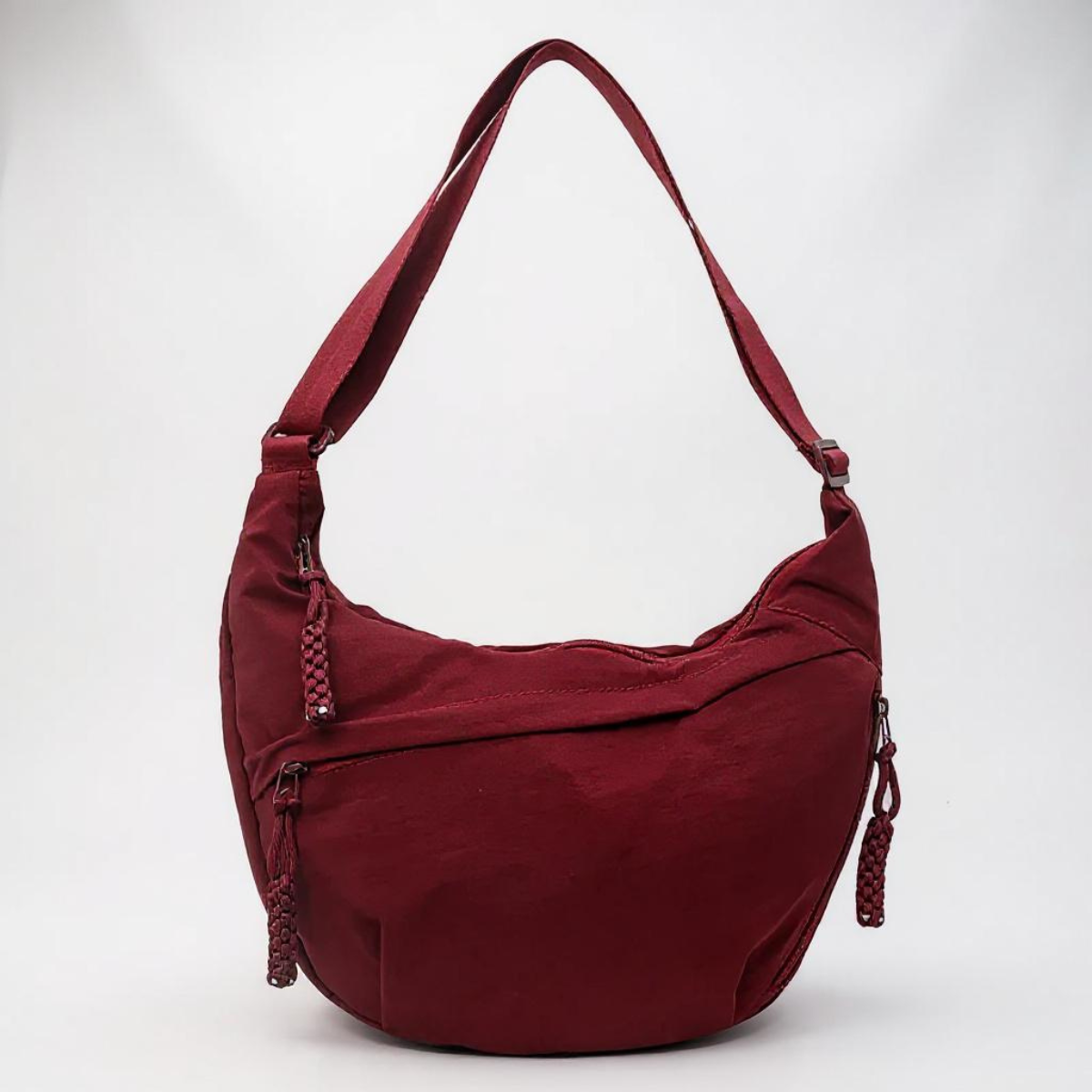 Halbmondförmige Schultertasche in Bordeaux mit schräg verlaufenden Reißverschlüssen, geflochtenen Zippern und aufgestelltem Tragegurt vor hellem Hintergrund. 1