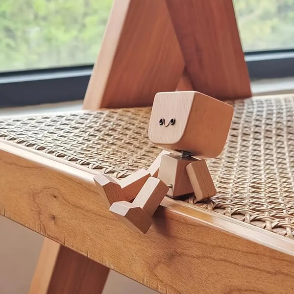 Holzfigur mit lächelndem Gesicht sitzt auf der Kante eines Holzstuhls mit Rattangeflecht vor einem Fenster. 11