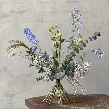Freistehender Strauß mit blauem Rittersporn, gelben Kugelblumen und Eukalyptus, unten an den Stielen zusammengehalten, vor grauer Wand. 5