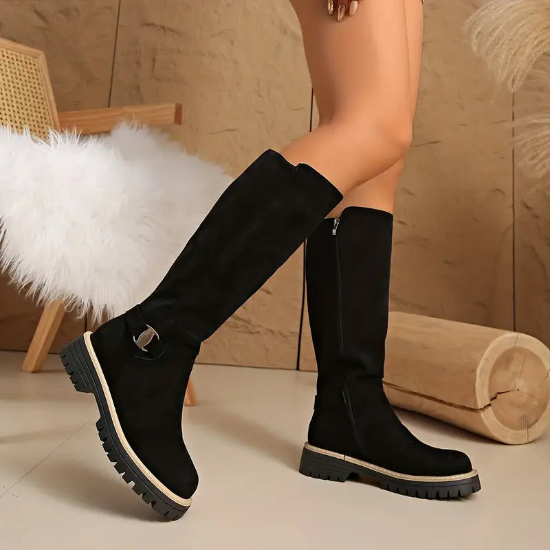 Schwarze hohe Stiefel aus Leder mit Metallschnalle und grober Profilsohle, getragen von einem Model vor Steinwand und Holzstamm.