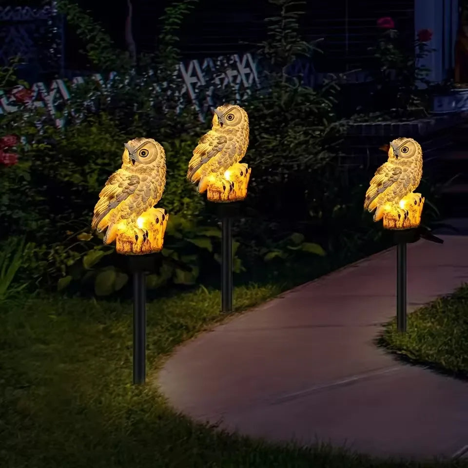 Drei gleich aussehende, warm leuchtende Eulen auf Spießen säumen abends einen geschwungenen Gartenweg. 9