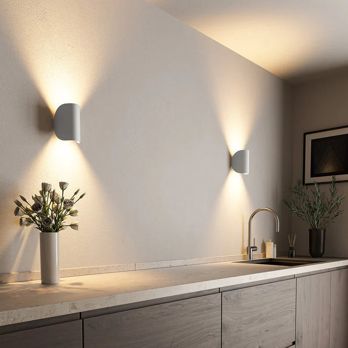Zwei weiße Wandleuchten mit Up-Down-Licht an einer Küchenwand neben Blumenvase, Spüle und gerahmtem Bild.