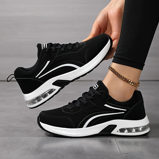 Schwarze Schnürsneaker mit weißer Luftpolstersohle, eine Hand hält einen Schuh angehoben, der andere Schuh steht auf grauem Boden. 1