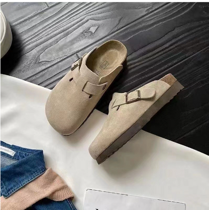 Graue Wildleder-Schlupfclogs mit Schnalle auf dunklem Holzboden, modische bequeme Schuhe.