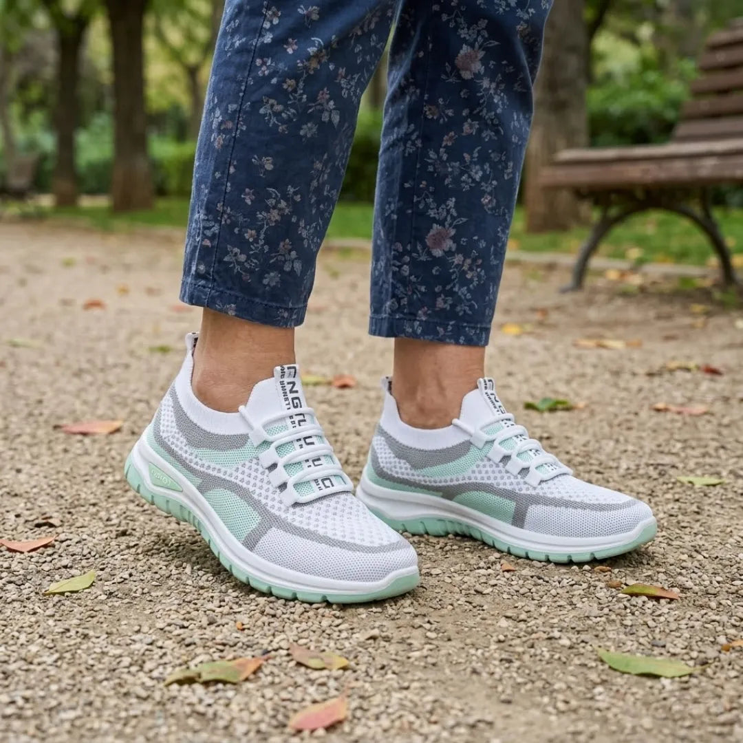 Weiß-mintgrüne Mesh-Sneaker an den Füßen einer Frau in geblümter Jeans auf Kiesweg vor einer Parkbank.