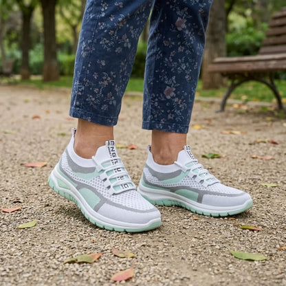 Weiß-mintgrüne Mesh-Sneaker an den Füßen einer Frau in geblümter Jeans auf Kiesweg vor einer Parkbank.