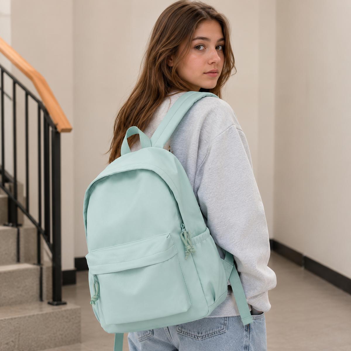 Junge Frau mit langen braunen Haaren trägt den mintgrünen Rucksack über der Schulter, im Treppenhaus mit hellgrauem Pullover und Jeans. 3