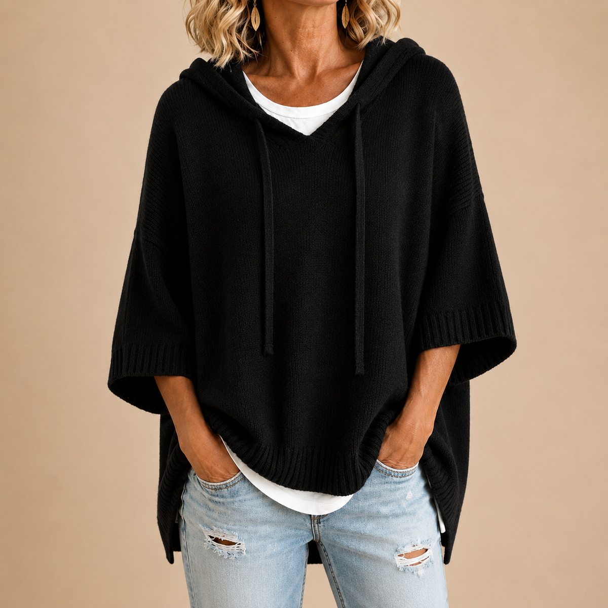 Schwarzer Strickponcho mit Kapuze und Kordeln, getragen mit weißem Shirt und hellen Jeans, Hände in den Hosentaschen, Vorderansicht. 1