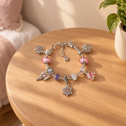 Fertig zusammengestelltes Charm-Armband mit rosa und lila Perlen sowie Schneeflocken- und Blumenanhängern auf einem runden Holztisch neben einer Zimmerpflanze. 1
