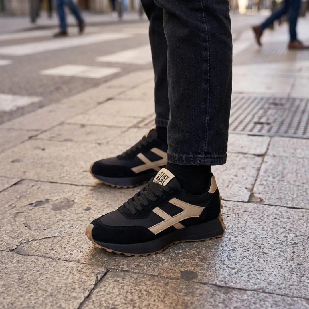 Schwarze Sneaker mit beigen Streifen, getragen zu dunkler Jeans auf Pflasterboden 17