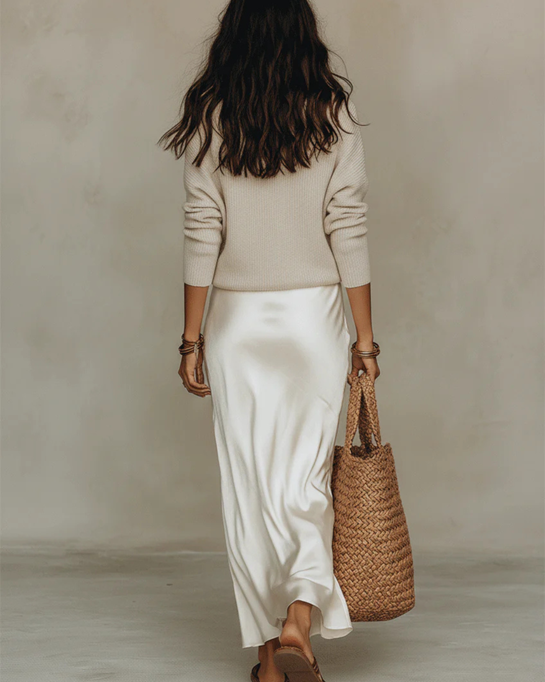 Rückansicht des Models in Creme: Strickpullover über langem Satinrock, dazu geflochtene Tasche und Sandalen 9
