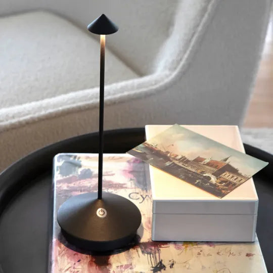 Schwarze, leuchtende Tischlampe mit Einschalttaste am Fuß auf einem Beistelltisch neben Bildband und weißer Box 22